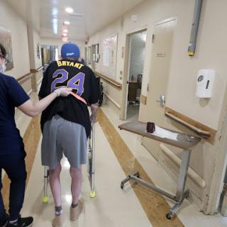 A caregiver assists a patient walking with a walker in a hospital corridor.