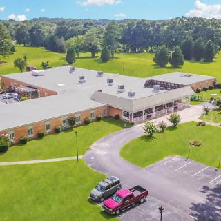 Aerial view of a large building surrounded by greenery with a parking lot and a few cars in front.