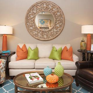 A stylish living room featuring a beige couch adorned with orange and green pillows, set against a decorative mirror. A glass coffee table displays vibrant vases, surrounded by modern lamps.
