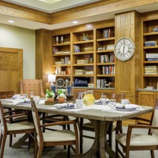 A cozy library meeting room with wooden bookshelves, a large table set for a gathering, and a wall clock accentuating the elegant interior.