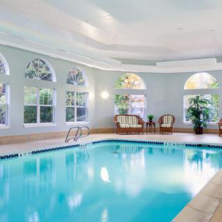 A serene indoor swimming pool area with bright lighting, surrounded by arched windows, chairs, and potted plants.