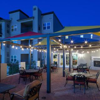 A cozy outdoor patio area adorned with string lights, featuring tables and chairs under a pergola, set against a modern building exterior during the evening.