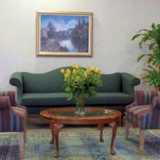 A cozy living room with a green sofa, flanked by colorful armchairs, and a wooden coffee table topped with a bouquet of yellow flowers. A framed painting hangs on the wall above.