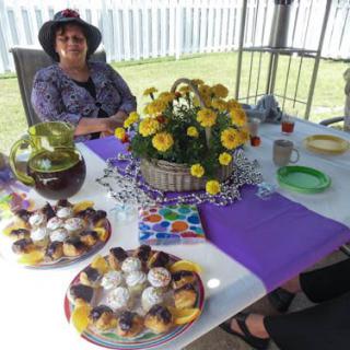 A person wearing a hat sits at a table decorated with flowers and desserts, set for a tea party outdoors.