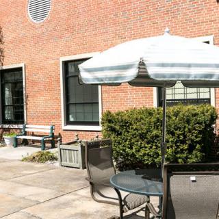 A sunny patio area with tables and chairs, featuring a large white umbrella and a backdrop of a brick building.