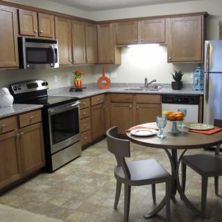 A modern kitchen with wood cabinets and stainless steel appliances, featuring a small dining area with a table and chairs.