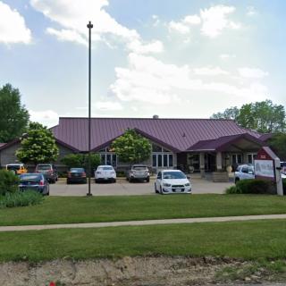 A single-story building with a parking lot in front, surrounded by trees and a grassy lawn under a partly cloudy sky.