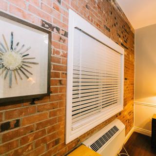 A cozy interior space featuring a brick wall, window with blinds, and a stylish clock above a modern air conditioner unit.
