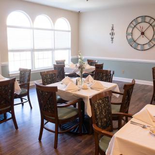 A cozy restaurant dining area with neatly arranged tables and chairs, featuring a large wall clock and soft natural light from arched windows.
