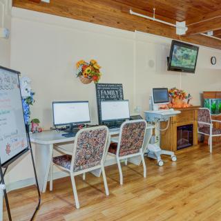 A cozy computer room with multiple workstations, featuring computers on desks, flower decorations, and a wall-mounted television.