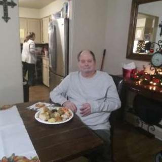 An elderly man enjoying a meal at the dining table while family members are busy in the kitchen.