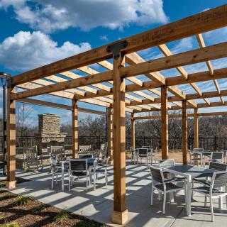 An outdoor patio with a wooden pergola, featuring tables and chairs under a clear, sunny sky.
