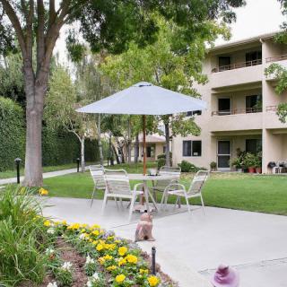 A serene outdoor patio area with a table, chairs, and umbrella set amidst a landscaped garden in front of an apartment building.