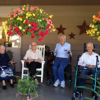 A group of elderly individuals sitting on a porch with hanging flower baskets and enjoying the outdoors.