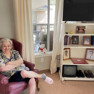 An elderly woman sitting comfortably in a living room next to a bookshelf with family photographs and a television.