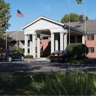 A two-story building with white columns and multiple flags flying at the entrance, surrounded by cars and landscaping on a sunny day.