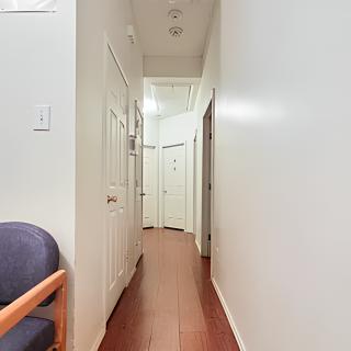A narrow hallway with wooden flooring and white walls, featuring multiple doors and a chair on the left side.