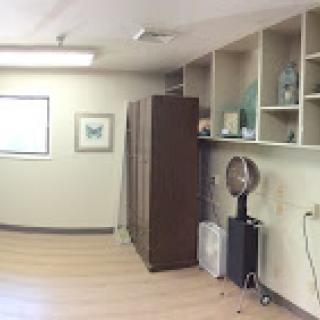 A spacious interior room featuring wooden flooring and various furniture pieces, including shelving and a fan.
