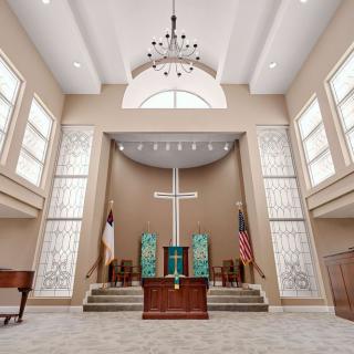 A church interior featuring an altar with a cross, surrounded by seating and large windows.