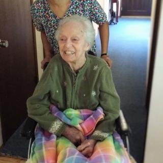 An elderly person sitting in a wheelchair, smiling warmly while covered with a colorful blanket indoors.