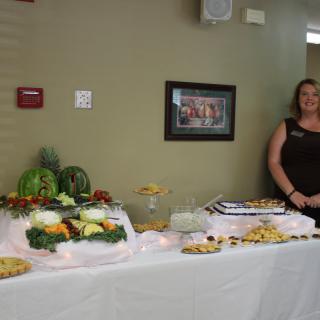 A woman stands beside a buffet table with a display of fruits, vegetables, and various dishes.