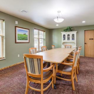 A well-lit conference room with a long wooden table surrounded by chairs, featuring a cabinet and windows.