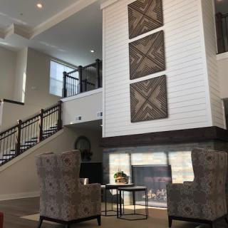 A modern living room interior with a high ceiling, featuring a staircase, artwork on the wall, and a cozy seating area by the fireplace.