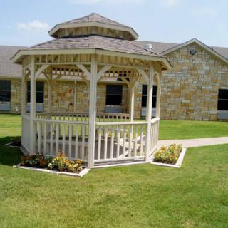 A charming white gazebo surrounded by well-maintained green grass and a stone building in the background.