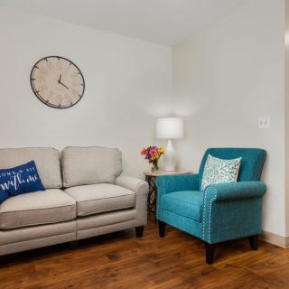 A cozy living room with a beige sofa and a blue armchair, accessorized with a wall clock and a side table lamp.