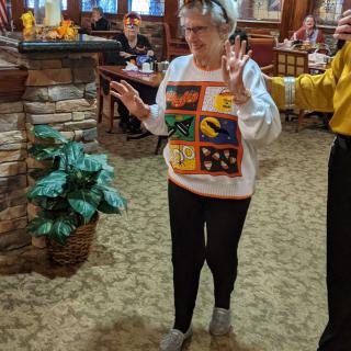 An elderly woman dances inside a restaurant, wearing a colorful sweater.