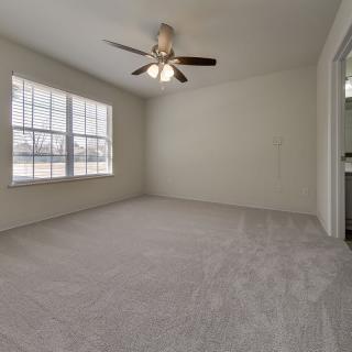 An empty room with carpet flooring, a ceiling fan, and large windows allowing natural light.