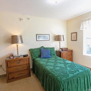 A cozy bedroom with a single bed featuring green bedding, flanked by wooden nightstands and lamps, with wall art and a window providing natural light.