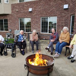 A group of elderly people sitting around a fire pit outside a brick building.