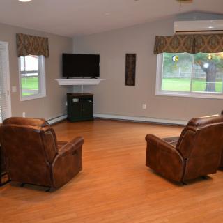 A minimalist living room with two leather chairs facing a wall-mounted television, featuring hardwood floors and large windows.