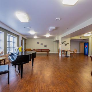 A spacious recreational room featuring a piano, foosball table, and seating area with large windows and hardwood floors.