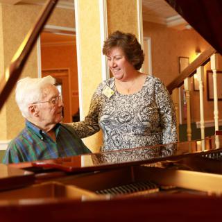 An elderly person and a woman are engaged in conversation near a piano in an indoor setting.
