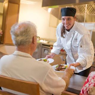 A chef serves a delicious meal to an elderly guest in a warm and welcoming kitchen environment.