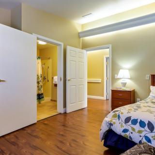 A cozy bedroom with wooden flooring, a bed with a patterned duvet, a lamp on the nightstand, and open doors leading to an adjoining room.