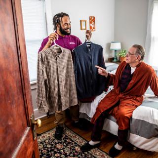 A caregiver presents clothing options to an elderly man, discussing fashion choices in a cozy bedroom setting.