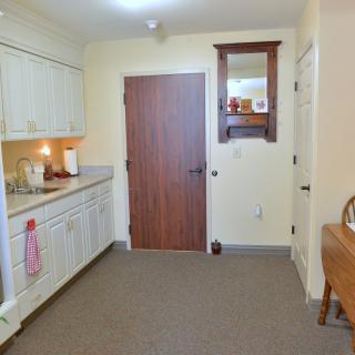 A cozy indoor kitchen with white cabinets, a sink, and a wooden door. The floor is covered with carpet.