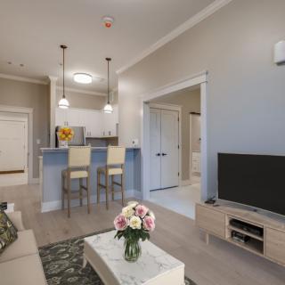 A modern living room and kitchen area featuring a TV, bar stools, and a vase of flowers on the coffee table.