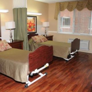 A nursing home or hospital room with two beds, separated by a curtain, featuring wooden furniture and a large window.