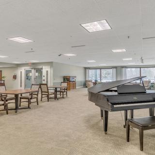 A spacious interior room featuring a grand piano and several tables with chairs, illuminated by natural light from the windows.