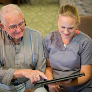 An elderly man and a nurse smiling while looking at a tablet together.
