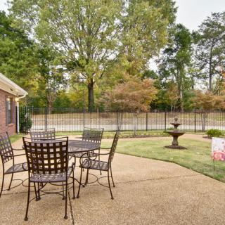 A tranquil patio area with a table and chairs overlooks a landscaped garden featuring a fountain and surrounded by trees.