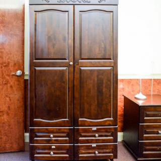 A wooden wardrobe with intricate carvings stands next to a matching drawer unit in a room.