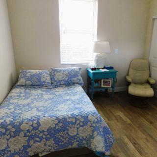 A cozy bedroom with a floral bedspread, a nightstand with a lamp, and a chair by the window.