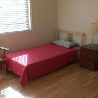 A minimalist bedroom with a single bed covered in red bedding, a wooden nightstand with a lamp, and a chair by the window.