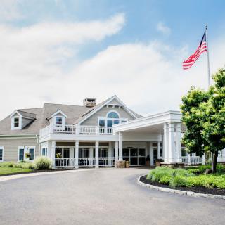 A large building with a sloping roof and a porch area, surrounded by greenery and an American flag flying on the right side.