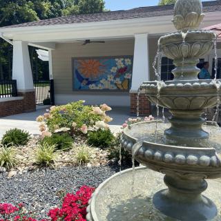 A serene garden setting featuring a tiered fountain, colorful flowers, and a building with outdoor artwork.